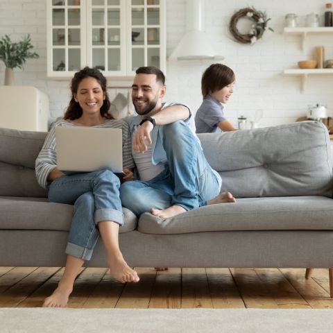 Family relaxes in the living room on a couch, while the parents work on the laptop and the children play. Bright, modern decor with wooden flooring and an open kitchen in the background.